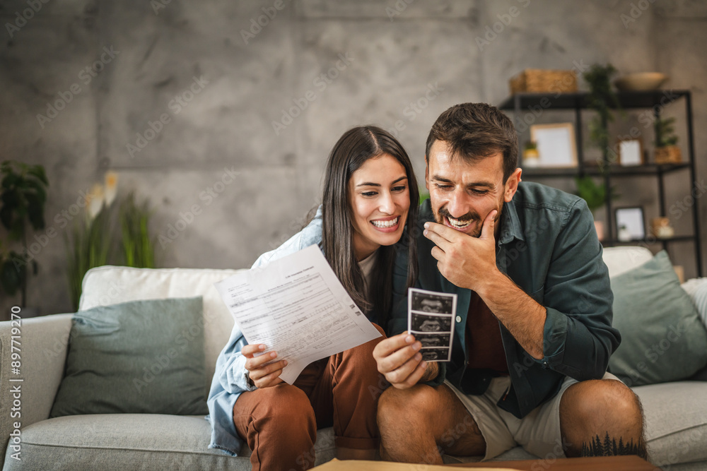 Adult couple sit on sofa and hold ultrasound picture of baby Adult couple sit on sofa and hold ultrasound picture of baby