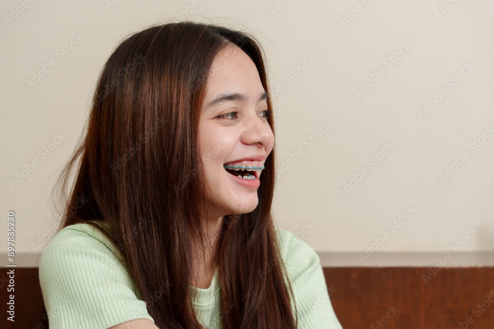 Asian teenage girl smiling brightly with braces. Wearing light green ribbed top, long straight brown hair. Warm indoor setting with beige walls and wooden panels. Expressing joy and happiness. Asian teenage girl smiling brightly with braces. Wearing light green ribbed top, long straight brown hair. Warm indoor setting with beige walls and wooden panels. Expressing joy and happiness.