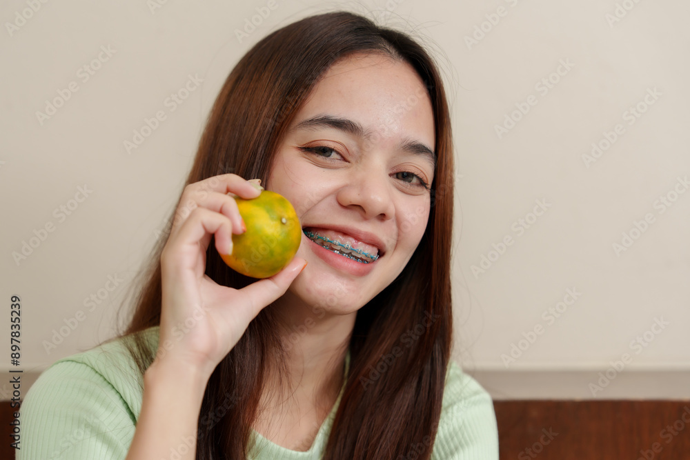 Young Asian girl with braces holding a fresh orange near cheek. Wearing light green ribbed top, long straight brown hair. Indoor setting with beige walls. Bright smile, showcasing dental braces, Young Asian girl with braces holding a fresh orange near cheek. Wearing light green ribbed top, long straight brown hair. Indoor setting with beige walls. Bright smile, showcasing dental braces,