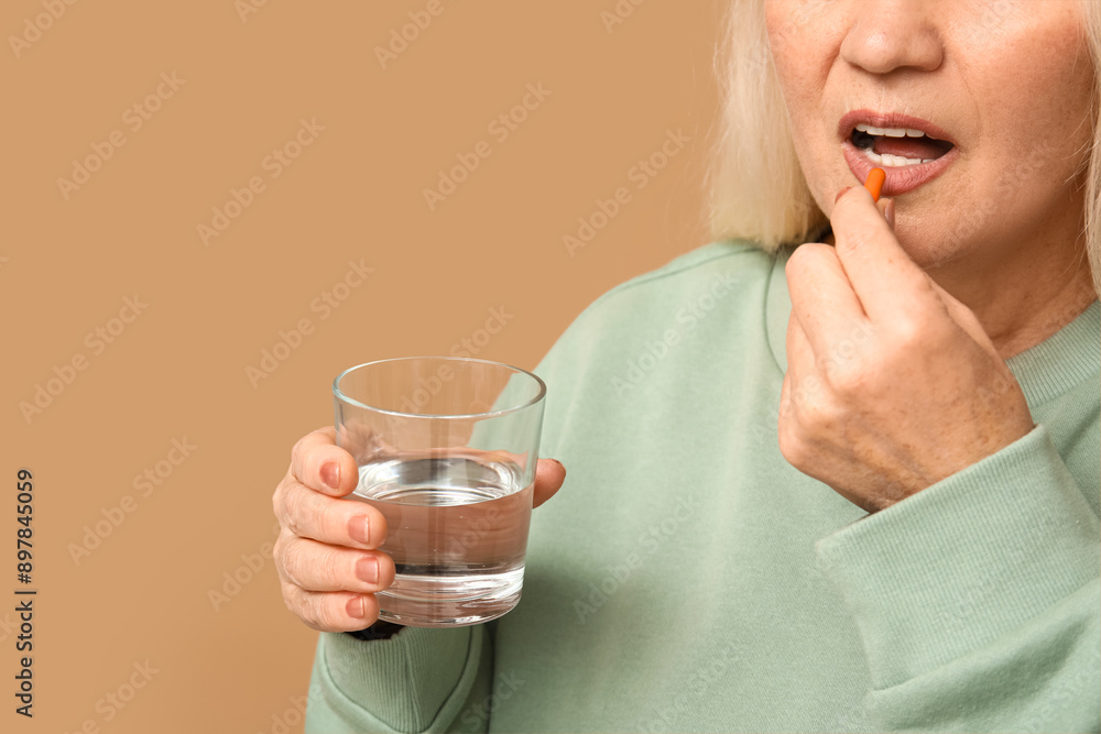 Beautiful mature woman with pill and glass of water on brown background Beautiful mature woman with pill and glass of water on brown background
