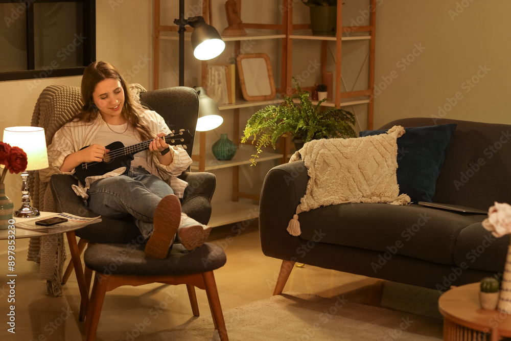 Obraz premium Beautiful young woman sitting in armchair and playing ukulele in evening