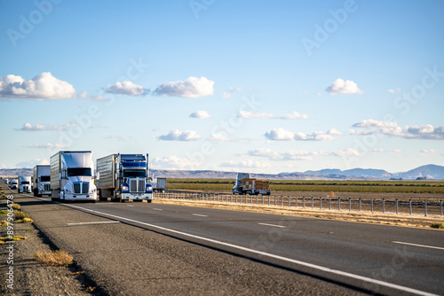 Convoy of the industrial big rig semi trucks with semi trailers transporting cargo running on the divided highway road in California