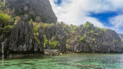 Picturesque karst rocks with steep slopes and sharp peaks surround the turquoise lagoon. Green tropical vegetation on the cliffs. Clouds in the blue sky. Philippines. Palawan.