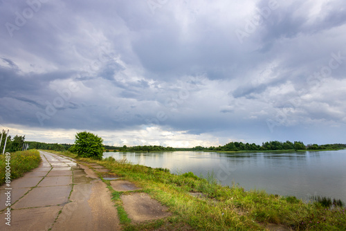 Wallpaper Mural A paved path winds through lush green grass and leads toward a calm river with a distant forest line. The sky is filled with dramatic, swirling clouds, suggesting an approaching storm.. Torontodigital.ca