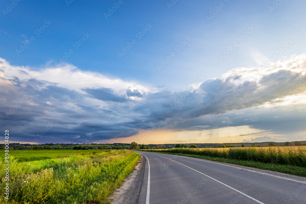 A long, paved road stretches through a rolling green landscape, with a dramatic, partly cloudy sky overhead. A long, paved road stretches through a rolling green landscape, with a dramatic, partly cloudy sky overhead.