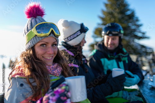 Friends enjoying a sunny winter day on the mountain, sipping tea and basking in the snowy landscape. They are bundled up in warm clothes, relishing the moment in the bright air