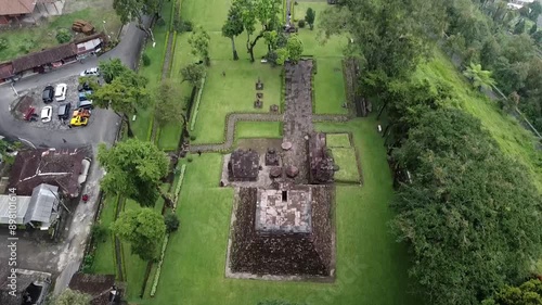 Sukuh Temple. Aerial view of an ancient temple complex surrounded by lush greenery. The temple features stone structures and pathways, with trees and a road visible nearby