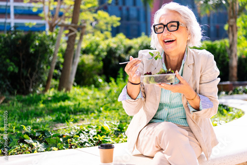 middle age entrepreneur woman laughing happy while eating a takeaway salad sitting outdoors in a city park, concept of healthy food and active lifestyle in adulthood, copyspace for text