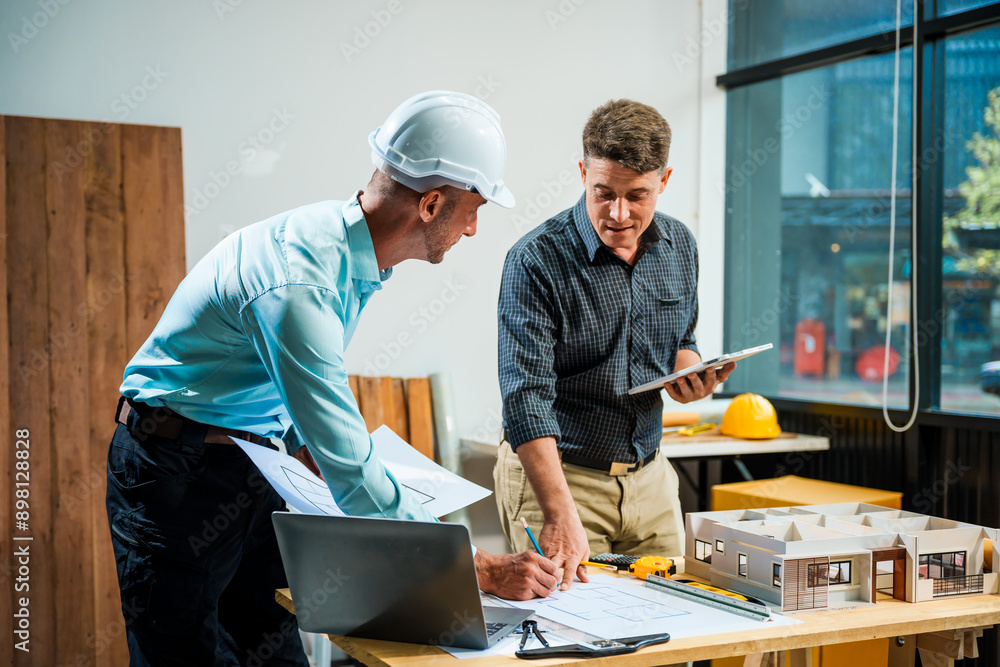A Caucasian male engineer and an Italian male building contractor are planning and talking at a desk, reviewing house plans and a model of a house with wooden textures for renovation. A Caucasian male engineer and an Italian male building contractor are planning and talking at a desk, reviewing house plans and a model of a house with wooden textures for renovation.