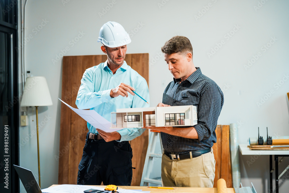 A Caucasian male engineer and an Italian male building contractor are planning and talking at a desk, reviewing house plans and a model of a house with wooden textures for renovation. A Caucasian male engineer and an Italian male building contractor are planning and talking at a desk, reviewing house plans and a model of a house with wooden textures for renovation.