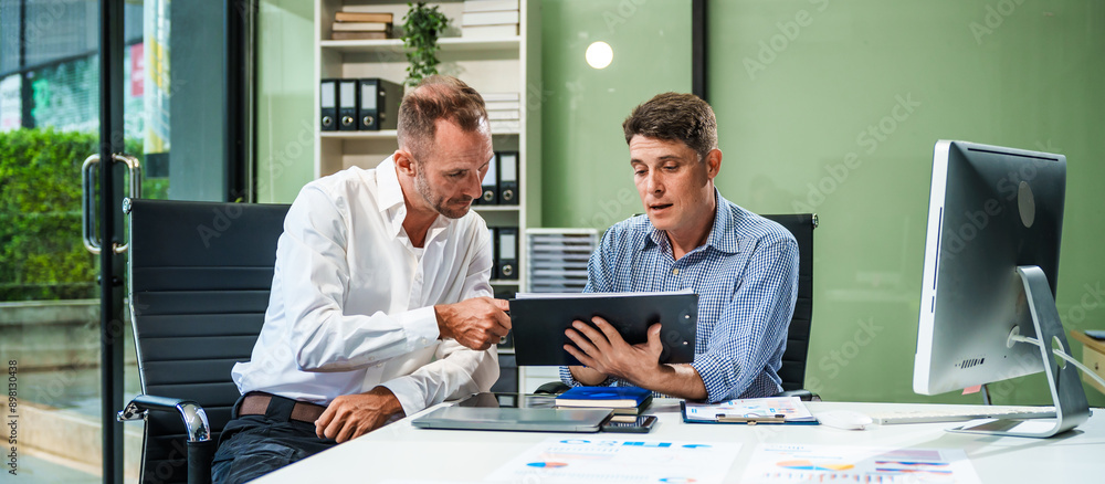 Fototapeta premium An Italian male and a Caucasian male sit at business desk in office meeting room, discussing financial terms, investment strategies, managing funds with computer monitor displaying relevant data.
