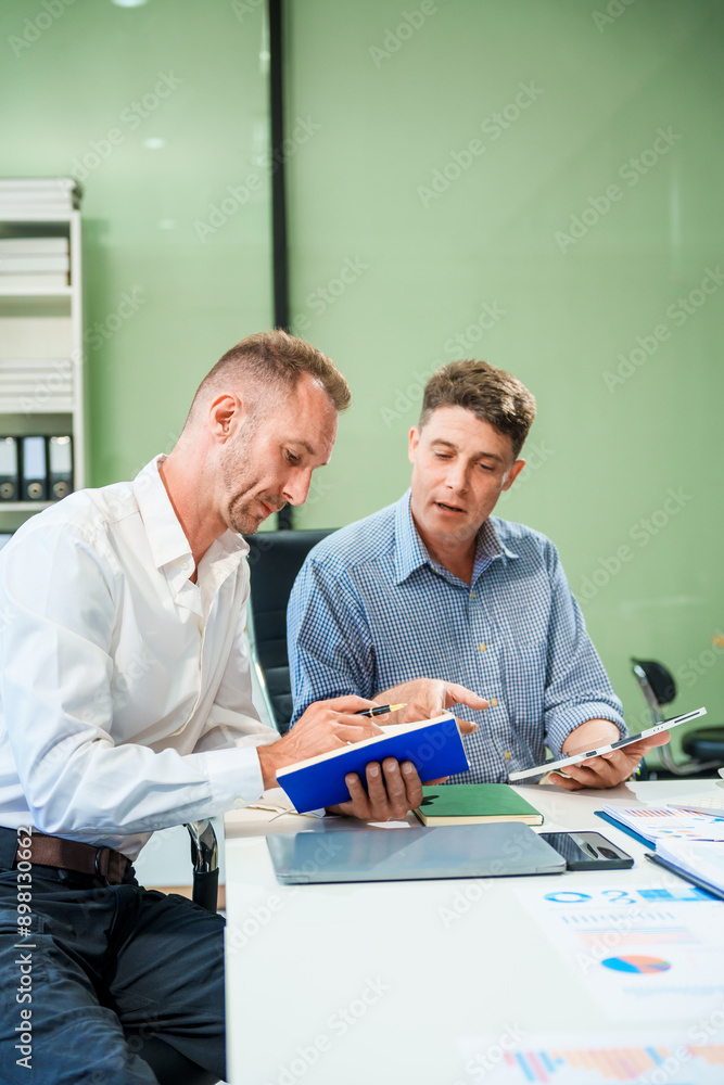 Obraz premium An Italian male and a Caucasian male sit at business desk in office meeting room, discussing financial terms, investment strategies, managing funds with computer monitor displaying relevant data.