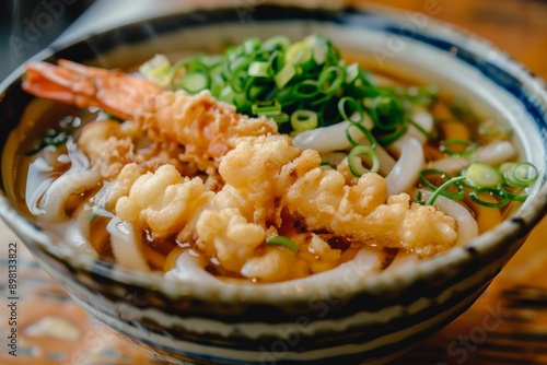 A steaming bowl of udon noodles topped with crispy tempura shrimp, garnished with spring onions and served in a traditional Japanese ceramic bowl on a wooden table. photo