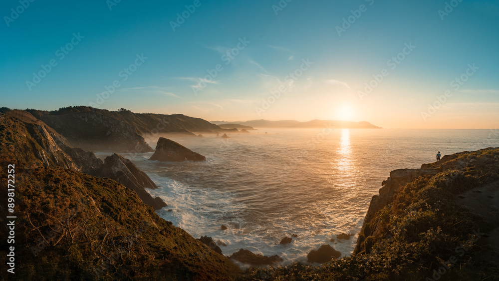 The Loiba cliffs at sunset with the sun casting a golden light. A person is seen in the distance admiring the view. The sky is filled with soft, wispy clouds. The Loiba cliffs at sunset with the sun casting a golden light. A person is seen in the distance admiring the view. The sky is filled with soft, wispy clouds.