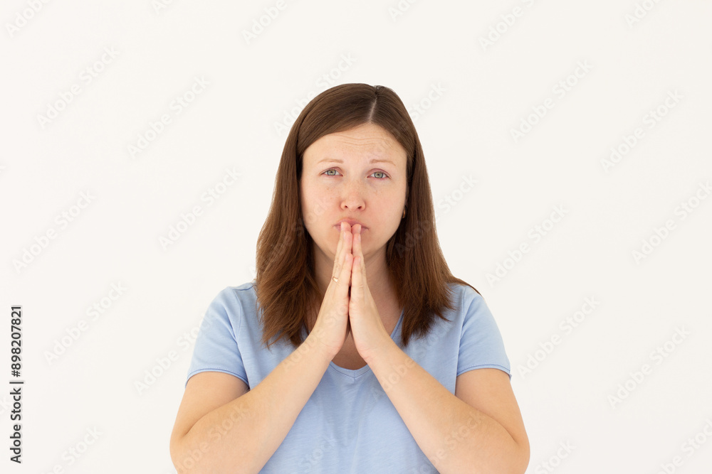 Portrait of imploring girl tilts head, purses lips, looks sadly at camera, makes praying gesture, asks for help and support, begs boyfriend for apologize, wears blue t-shirt, isolated on white wall Portrait of imploring girl tilts head, purses lips, looks sadly at camera, makes praying gesture, asks for help and support, begs boyfriend for apologize, wears blue t-shirt, isolated on white wall