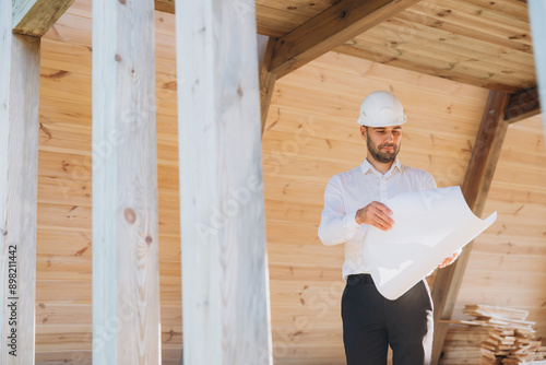 Construction engineer in white helmet. Developer with construction documentation on the background of wooden modular building under construction. Portrait of smiling architect near eco friendly house.