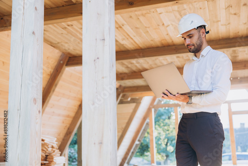 Man builder. Foreman with laptop. Contractor for construction company. Architect in unfinished building. Builder in wooden modular house under construction. Man engineer from construction team © anatoliycherkas