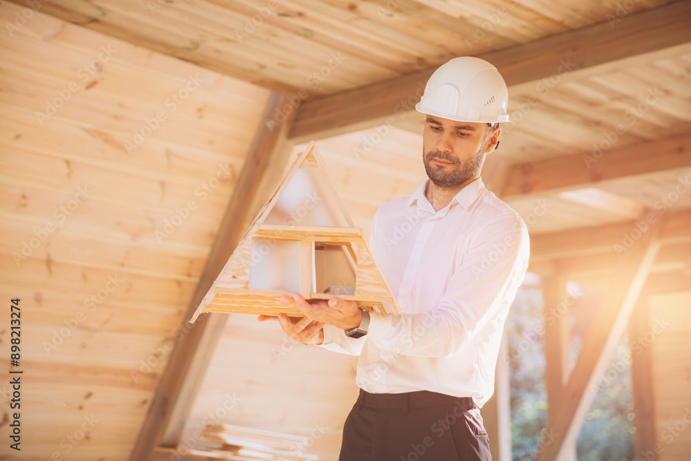 Man builder. Contractor for construction company. Architect in unfinished building. Builder in wooden modular house under construction. Male engineer holding a model of a miniature wooden house Man builder. Contractor for construction company. Architect in unfinished building. Builder in wooden modular house under construction. Male engineer holding a model of a miniature wooden house