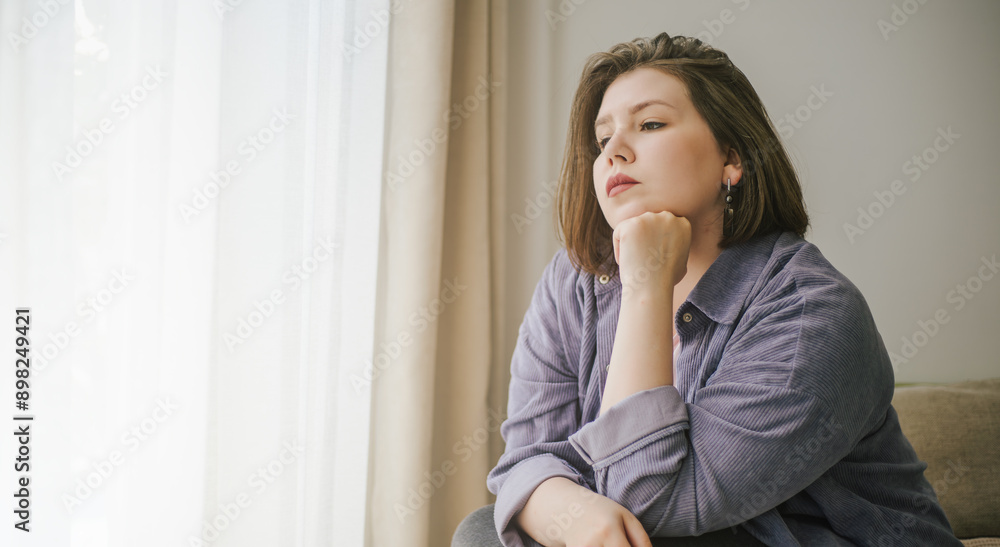 Plus size woman sits indoors, gazing thoughtfully out window alone, lost in contemplation. Plus size woman sits indoors, gazing thoughtfully out window alone, lost in contemplation.