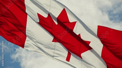 A vibrant Canadian flag waves proudly in the wind against a clear blue sky with fluffy white clouds and a bright sun. Slow motion.