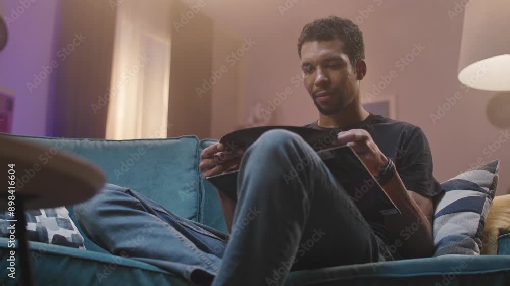 Young African American guy holding vinyl record while sitting on soft couch in dim living room. Handsome man considering musical plate while resting at home alone. Old music. Vintage. Young African American guy holding vinyl record while sitting on soft couch in dim living room. Handsome man considering musical plate while resting at home alone. Old music. Vintage.