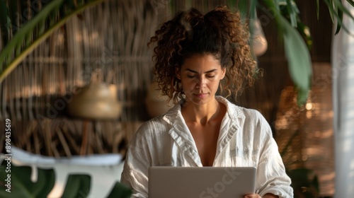 A focused woman with curly hair wears a white blouse as she works on her laptop in a natural, serene environment, surrounded by supportive greenery and soft light. photo