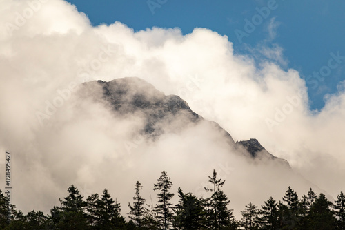 mountain scene with a dramatic interplay of light and clouds. A majestic mountain peak emerges from a thick layer of white clouds, partially veiled in mist. 