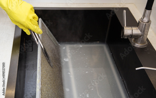 A girl's hand in a yellow glove turns over a filter from a kitchen hood with tongs in the cleaning mixture of an oxygen purifier for the kitchen. Chemical reaction