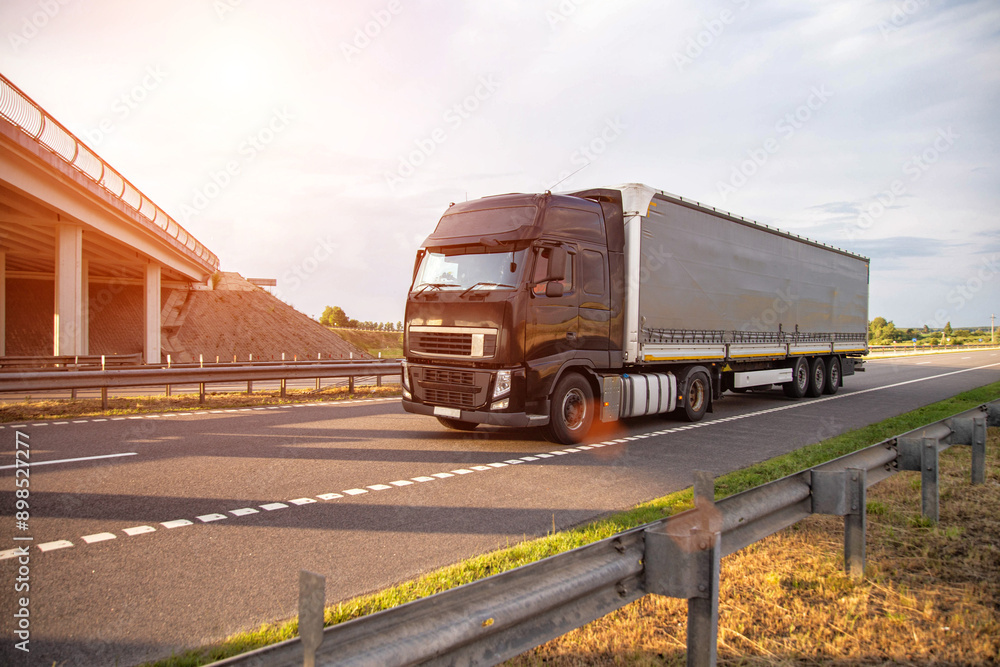a black modern truck with a semi-trailer is driving along the road against the backdrop of a bridge and sunset. Freight transportation services, business. Copy space for text a black modern truck with a semi-trailer is driving along the road against the backdrop of a bridge and sunset. Freight transportation services, business. Copy space for text