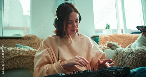 Happy brunette girl in black headphones is engaged in her hobby and adjusts music using a DJ mixer at home near the sofa in a modern apartment