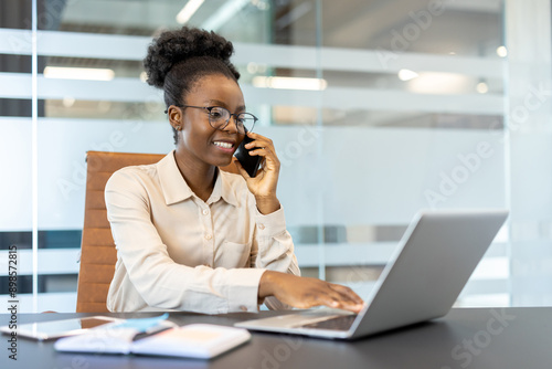Smiling businesswoman in glasses talking on phone while working on laptop in modern office. Image portrays professionalism, productivity, and modern workplace environment.