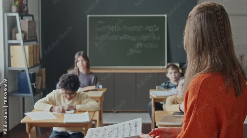 Young female teacher holding sheet music and asking question during lesson in primary school, all kids raising their hands, African American boy answering Young female teacher holding sheet music and asking question during lesson in primary school, all kids raising their hands, African American boy answering