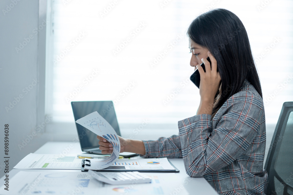 A woman is talking on her cell phone while sitting at a desk with a stack of pap A woman is talking on her cell phone while sitting at a desk with a stack of pap