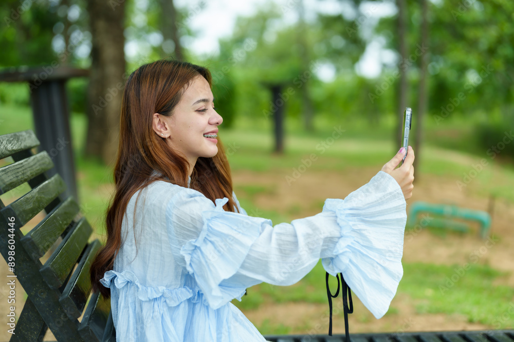 Naklejka premium Asian woman in blue blouse and white pants sitting on black bench, taking a selfie with phone, smiling. Background includes trees and grass in park, capturing cheerful, fun moment outdoors