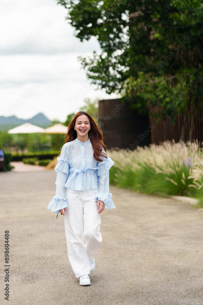 Young Asian woman in blue blouse and white pants walking on a pathway, smiling broadly. Background includes trees, greenery, and distant mountains. Captures joyful moment during outdoor stroll.