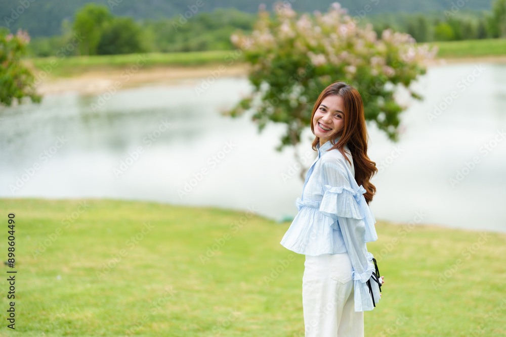 Asian woman in blue blouse and white pants standing by a lake, smiling. Background shows green grass, blooming trees, and calm water. Captures joyful, serene moment in nature. Asian woman in blue blouse and white pants standing by a lake, smiling. Background shows green grass, blooming trees, and calm water. Captures joyful, serene moment in nature.