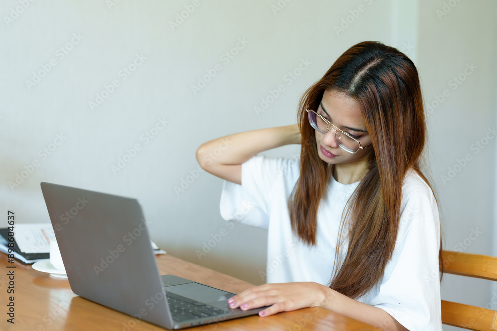 Asian woman in white shirt and glasses stretching neck while working on laptop at wooden table. Background includes a coffee cup. Captures a moment of relief during remote work Asian woman in white shirt and glasses stretching neck while working on laptop at wooden table. Background includes a coffee cup. Captures a moment of relief during remote work
