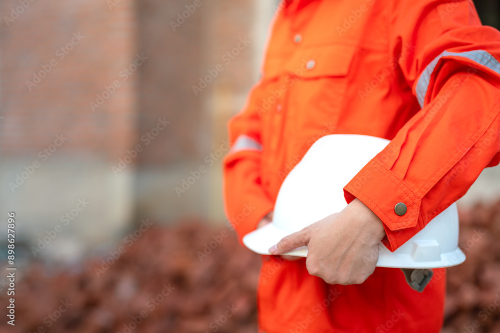An engineer or foreman in orange coverall uniform is holding a white safety helmet or hardhat, standing in front of construction building site. Safety work practice in danger workplace concept. An engineer or foreman in orange coverall uniform is holding a white safety helmet or hardhat, standing in front of construction building site. Safety work practice in danger workplace concept.