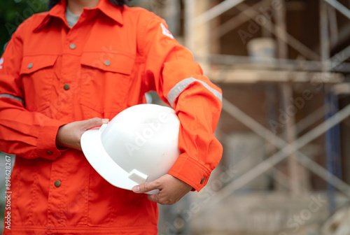 Fotografie An engineer or foreman in orange coverall uniform is holding a white safety helmet or hardhat, standing in front of construction building site