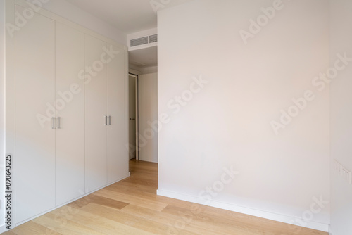 A minimalist bedroom featuring a built-in wardrobe with white doors, natural wood floor, and a wall ventilation system creating a modern and clean look.