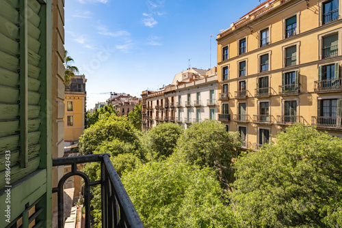 Daytime view of urban buildings adorned with balconies and surrounded by green leafy trees under a sunny sky, showcasing a harmonious blend of nature and architecture in Barcelona Spain