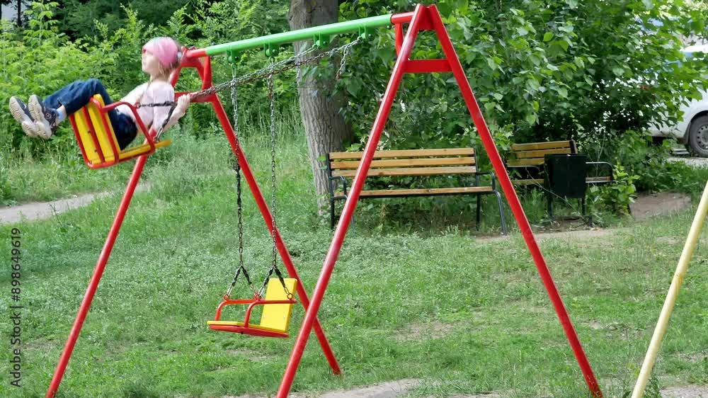 A little girl in jeans is quickly swinging on a swing on an outdoor playground in summer, green trees. A little girl in jeans is quickly swinging on a swing on an outdoor playground in summer, green trees.