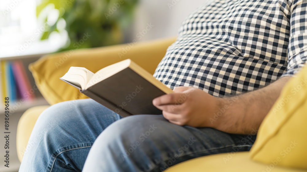 Man reading book while sitting on sofa, close-up of belly, casual attire, relaxed atmosphere, home comfort concept Man reading book while sitting on sofa, close-up of belly, casual attire, relaxed atmosphere, home comfort concept