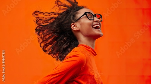 A joyful girl with black glasses, dressed in an orange outfit, is jumping with excitement and laughing. The bright orange background complements her lively and enthusiastic personality