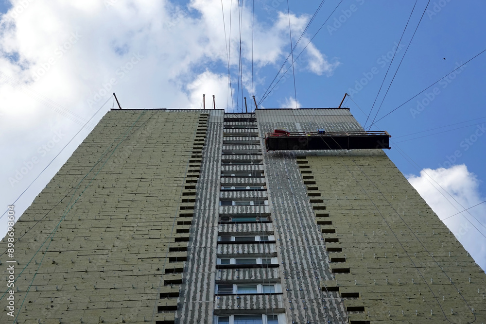 Obraz premium Bottom up view of being constructed high building with insulation on facade and construction cradle under sky with soft white clouds