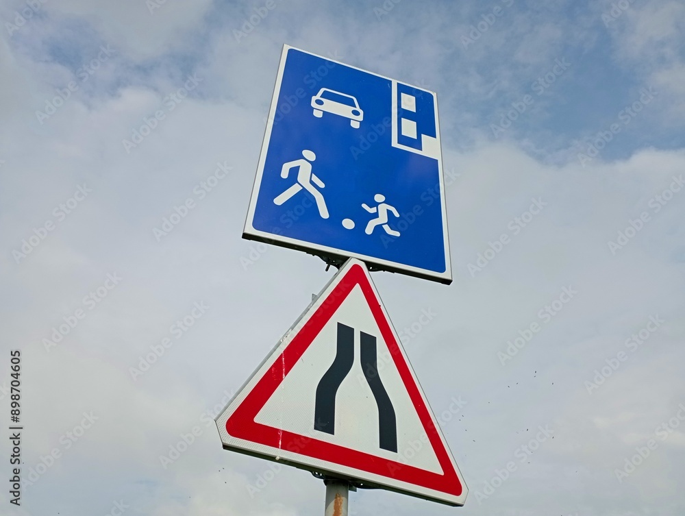 A triangular road sign narrowing the road above which is a warning sign about the place for walking and playing with children. A triangular road sign narrowing the road above which is a warning sign about the place for walking and playing with children.