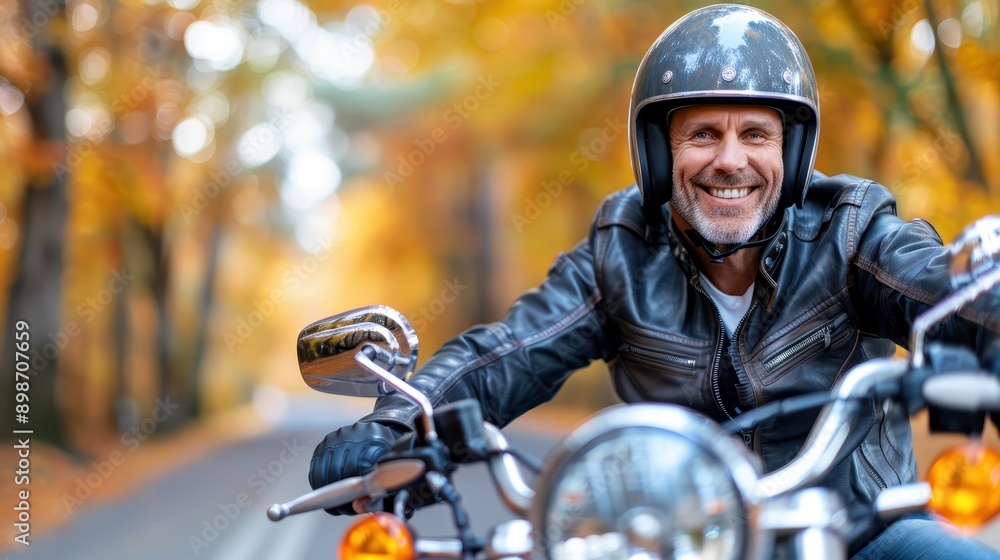 Fototapeta premium A smiling man wearing a black leather jacket and helmet rides his motorcycle down a wooded road, surrounded by the warm, vibrant colors of autumn leaves.