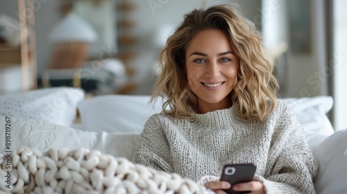 A woman with wavy hair smiles warmly on her bed, wrapped in a fuzzy knit blanket, exuding comfort, relaxation, and happiness in a serene bedroom setting. photo