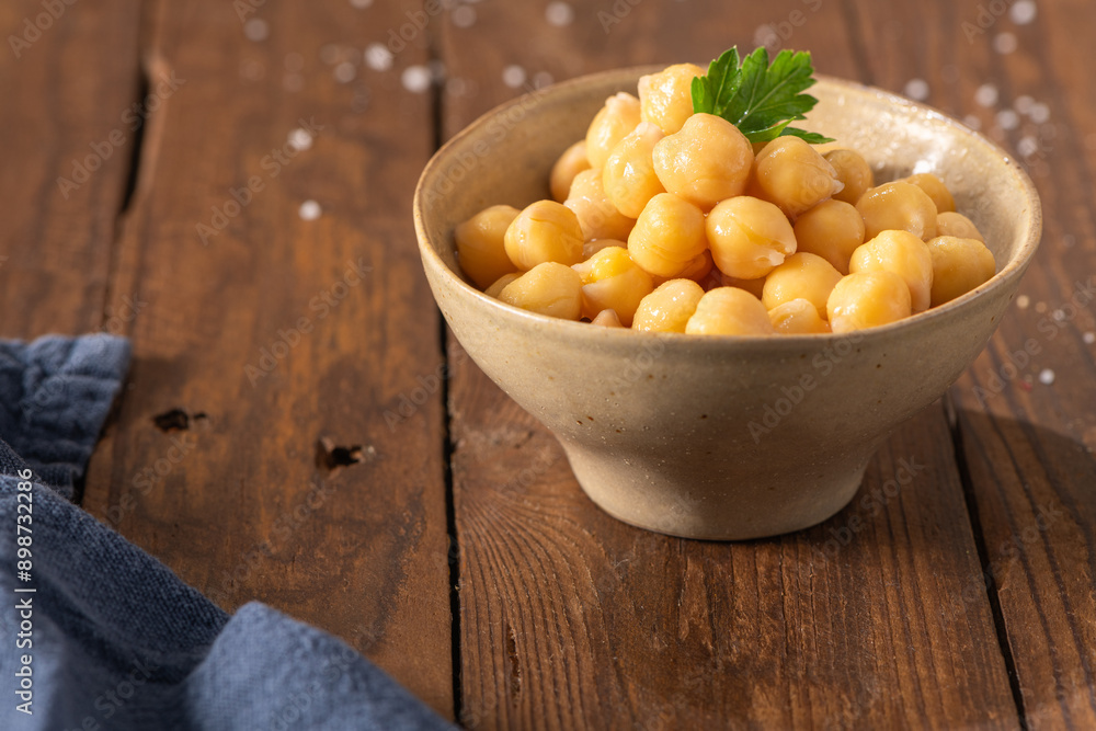 Cooked chickpeas in a ceramic bowl on a rustic wooden table decorated with a parsley leaf, ready to eat Cooked chickpeas in a ceramic bowl on a rustic wooden table decorated with a parsley leaf, ready to eat