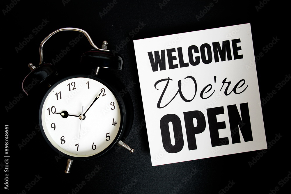 Welcome We're OPEN text with alarm clock on black background Welcome We're OPEN text with alarm clock on black background